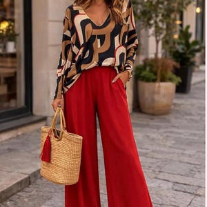 Könnte beinhalten: Ein Outfit mit einem langärmeligen Oberteil mit geometrischem Muster in Schwarz, Braun und Beige, kombiniert mit einer roten Hose mit weitem Bein. Eine gewebte Stroh-Tragetasche mit roter Quaste und Sonnenbrille vervollständigen den Look.