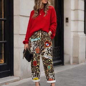 May include: A bright red long-sleeved jumper paired with leopard print trousers featuring various colourful patches including a heart with an eye, a rose, a lightning bolt, a smiley face, and the text XOXO and Stick With It, worn with black heels.