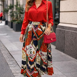 Pode incluir: Uma mulher com uma camisa de seda vermelha e uma saia midi plissada com um padrão geométrico e floral colorido. Ela carrega uma bolsa vermelha com uma corrente dourada e saltos vermelhos. O conjunto é complementado com joias douradas e óculos de sol.
