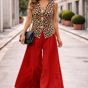 May include: A woman in a sleeveless leopard print top with cherry detailing and gold buttons, teamed with wide-leg red trousers. She is also wearing black sunglasses, gold jewellery, and carrying a black quilted handbag.
