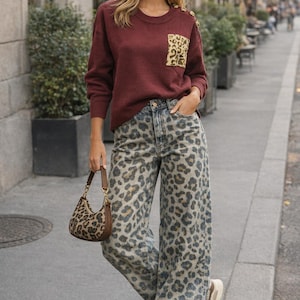Könnte beinhalten: Ein Outfit für Damen mit einem weinroten Pullover mit Leopardenmuster-Tasche und goldenen Knöpfen, kombiniert mit Jeans mit Leopardenmuster und einer passenden Handtasche. Das Outfit wird durch beige Sneaker ergänzt.