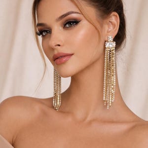 Könnte beinhalten: Elegante goldfarbene Ohrringe mit einem rechteckigen Kristall-Stecker und mehreren Strängen aus kleinen, funkelnden Kristallen. Die Ohrringe fallen herab und erzeugen einen glamourösen, auffälligen Effekt. Das Bild zeigt das filigrane Design und die Lichtreflexion.