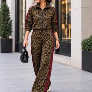 Puede incluir: Un conjunto de dos piezas a juego que incluye una chaqueta de chándal con cremallera y pantalones de pierna ancha con estampado de leopardo verde oliva. Ambas piezas tienen rayas laterales de color rojo oscuro en mangas y piernas. Bolso negro.