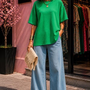 Könnte beinhalten: Ein leuchtend grünes Kurzarm-Oberteil mit dezenter horizontaler Rippstruktur, kombiniert mit einer hellblauen, weit geschnittenen Jeans. Das Outfit wird durch eine beige geflochtene Clutch mit langen Fransen sowie eine goldene Kette und Armspange ergänzt.