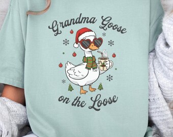 May include: A light green t-shirt featuring a cartoon goose wearing sunglasses, a Santa hat, and a scarf, holding a mug. The text "Grandma Goose on the Loose" is above and below the goose illustration. The shirt is a relaxed fit.