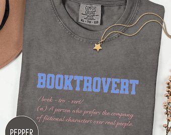 May include: A gray Comfort Colors t-shirt with the word "BOOKTROVERT" in blue and a definition in pink. A gold star pendant necklace rests on the shirt. The shirt is size XL.