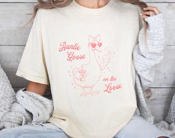 Może przedstawiać: Kremowa koszulka z różową grafiką gęsi w okularach przeciwsłonecznych w kształcie serca i trzymającej napój. Tekst "Auntie Goose on the Loose" znajduje się nad i pod ilustracją gęsi. Koszulka ma krótkie rękawy i luźny krój.
