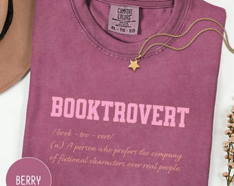 May include: Berry-colored t-shirt with the word "BOOKTROVERT" in pink block letters. The definition of "booktrovert" is printed below. A gold star pendant necklace rests on the shirt. The shirt is size XL.