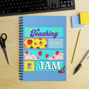 May include: A blue spiral-bound notebook featuring the phrase "Teaching is my JAM" in a colourful, comic-book style. The design incorporates strawberries, clouds, and a jam jar. A black keyboard, scissors, and pens are in the background.