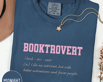 May include: A navy blue t-shirt with the word "BOOKTROVERT" in pink block letters. The definition of "booktrovert" is printed below in a smaller font. A gold star pendant necklace rests on the shirt.