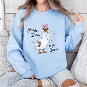 Könnte beinhalten: Hellblaues Sweatshirt mit einem Cartoon-Gans-Design, das eine Sonnenbrille und eine rosa Schleife trägt und einen Kaffeebecher hält. Der Text lautet "Auntie Goose on the Loose". Das Sweatshirt hat einen Rundhalsausschnitt.