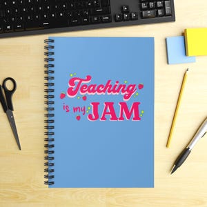 Puede incluir: Un cuaderno de espiral azul claro con el texto "Teaching is my JAM" en rosa, decorado con fresas y estrellas. También se ven un teclado, tijeras, un lápiz y un bolígrafo.