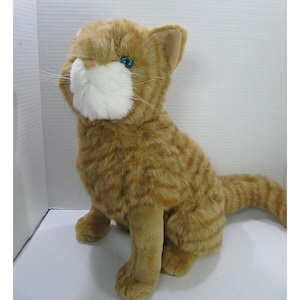 May include: A plush orange tabby cat with green eyes and white whiskers. The stuffed animal is sitting upright with its tail extended. The cat has a white muzzle and a soft, furry texture.