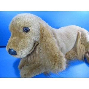 May include: A plush, golden-brown Cocker Spaniel dog toy with a black nose and dark eyes. The toy has long, floppy ears and a soft, furry texture. The dog is lying down on a blue surface.