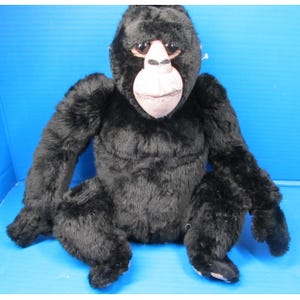 May include: A plush gorilla toy featuring soft black fur covering its body, long arms, and a light-colored face with dark eyes. The stuffed animal is depicted in a seated position with its limbs extended outward, showcasing a realistic design.