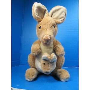 May include: A plush kangaroo toy in shades of brown and tan. The kangaroo has large ears, a detailed face, and a pouch with a smaller joey. The toy is made of soft, textured material.