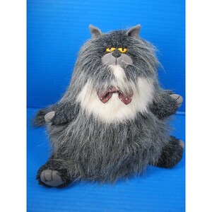 May include: A plush cat toy with gray fur, a white chest, and a maroon bow tie with white polka dots. The cat has yellow eyes and gray paws. The toy is sitting down and has a long, shaggy appearance.