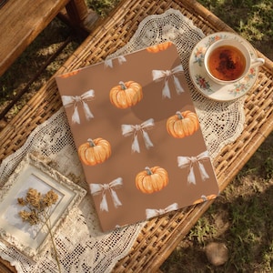 Pode incluir: Um caderno castanho com um padrão de abóboras laranja e laços brancos. Está sobre uma superfície de vime, ao lado de uma chávena de chá e de uma moldura com flores secas. A cena evoca um ambiente outonal acolhedor.