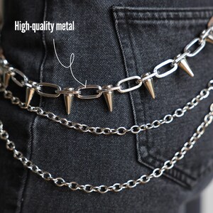 May include: Silver metal chain belt with three layers of chains and cone-shaped spikes. The text "High-quality metal" is visible in the image. The belt is worn over dark denim jeans.
