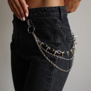 May include: Silver chain belt with multiple strands and a carabiner clip. The belt features a series of silver chains, a chain with large links, and a chain with gold-colored spikes. The belt is attached to black denim jeans.