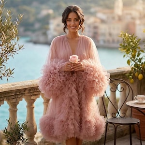 May include: A dusty rose tulle robe featuring a deep V-neckline, long voluminous sleeves with dense ruffled layers, and a matching ruffled hemline. The sheer fabric is layered throughout the garment, creating a textured and full appearance.