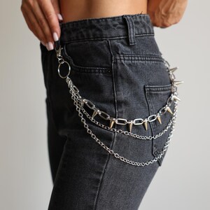 May include: Silver-toned chain belt with multiple strands and spiked accents. The belt is attached to a pair of dark gray denim jeans. The chain has a lobster clasp and a large ring.