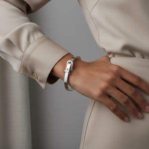 May include: A silver-colored bangle bracelet with a buckle design. The bracelet is worn on a wrist, and the buckle is centered on the front. The bracelet is made of a shiny metal and has a smooth finish. The background is a neutral color.