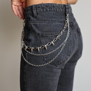 May include: Silver chain belt with three strands, featuring a row of silver spikes on the top chain. The belt is attached to the back pockets of dark gray denim jeans. The chains hang down, creating a layered effect.