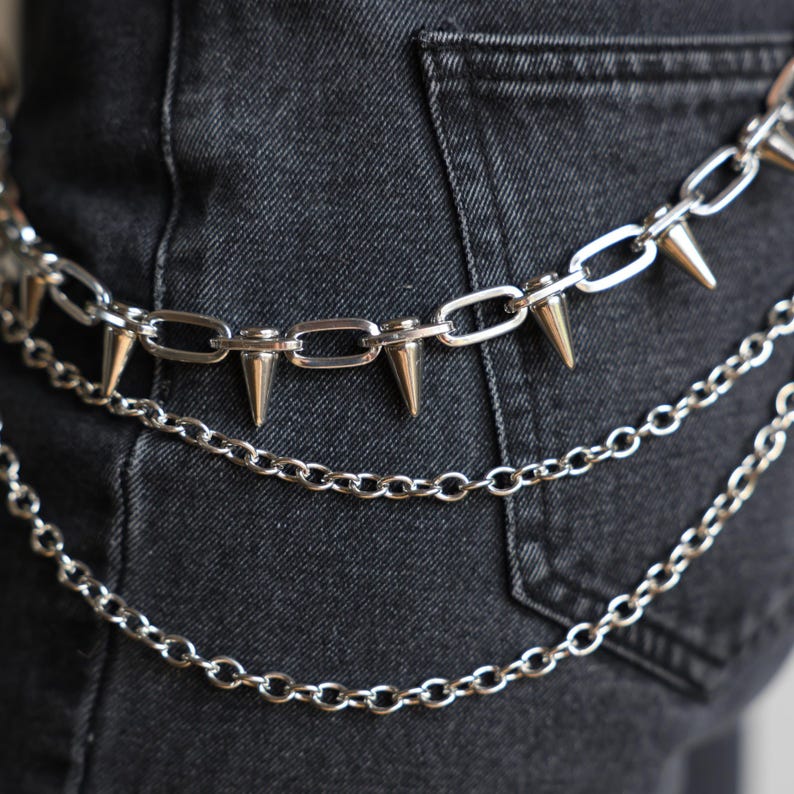 May include: Silver metal chain belt with three layers. The top layer features large, pointed spikes. The belt is shown against a dark denim background, highlighting the punk rock aesthetic. The chains are connected with oval links.
