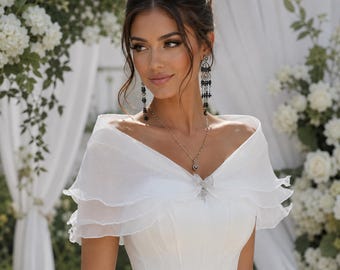 May include: A white wedding dress with a layered, off-the-shoulder cape. The dress features a fitted bodice and a flowing skirt. The model is wearing a necklace and earrings with black and silver accents. The background includes white flowers and drapery.