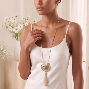 May include: A long gold-tone necklace featuring a large, textured shell-shaped pendant suspended from a thin chain. A cream-colored fabric tassel hangs directly from the bottom of the metallic shell pendant, creating a layered, vertical design.