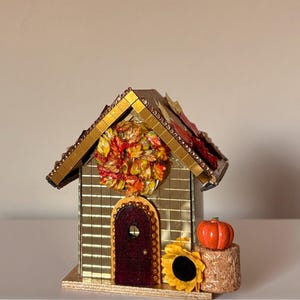 Pode incluir: Uma casa em miniatura decorativa coberta com azulejos de espelho dourados e uma porta em arco vermelho escuro. O telhado apresenta detalhes de folhas em tons de outono. Uma coroa de folhas de outono pende sobre a porta, junto a um girassol.