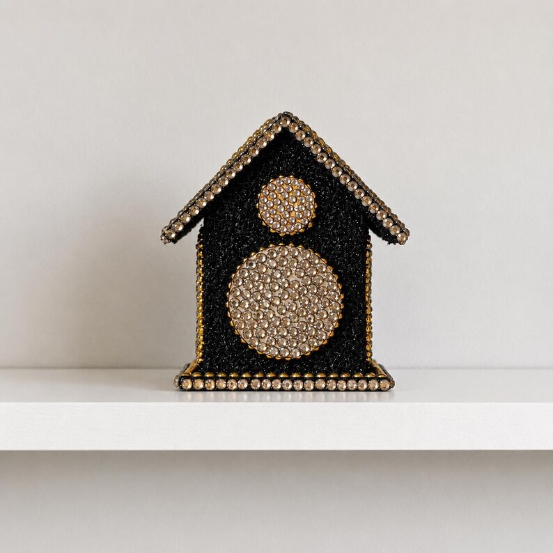 Black glitter decorative house with gold trim and sparkling crystal details, featuring two round rhinestone accents on the front, displayed alone on a clean white shelf against a soft neutral background.