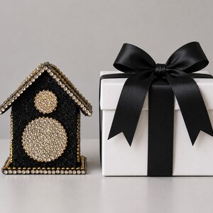 Black glitter and rhinestone keepsake house with gold accents displayed beside an white gift box tied with a satin bow.