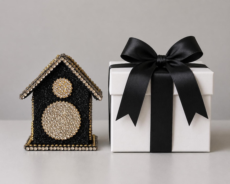 Black glitter and rhinestone keepsake house with gold accents displayed beside an white gift box tied with a satin bow.