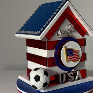 K&ouml;nnte beinhalten: Ein dekoratives patriotisches Haus mit blauem Dach, rot-wei&szlig;en W&auml;nden und einem Fu&szlig;ball. Das Haus zeigt ein USA-Flaggenemblem und das Wort "USA" in silbernen Buchstaben. Ein Fu&szlig;ballschuh und ein Ball sind ebenfalls abgebildet.