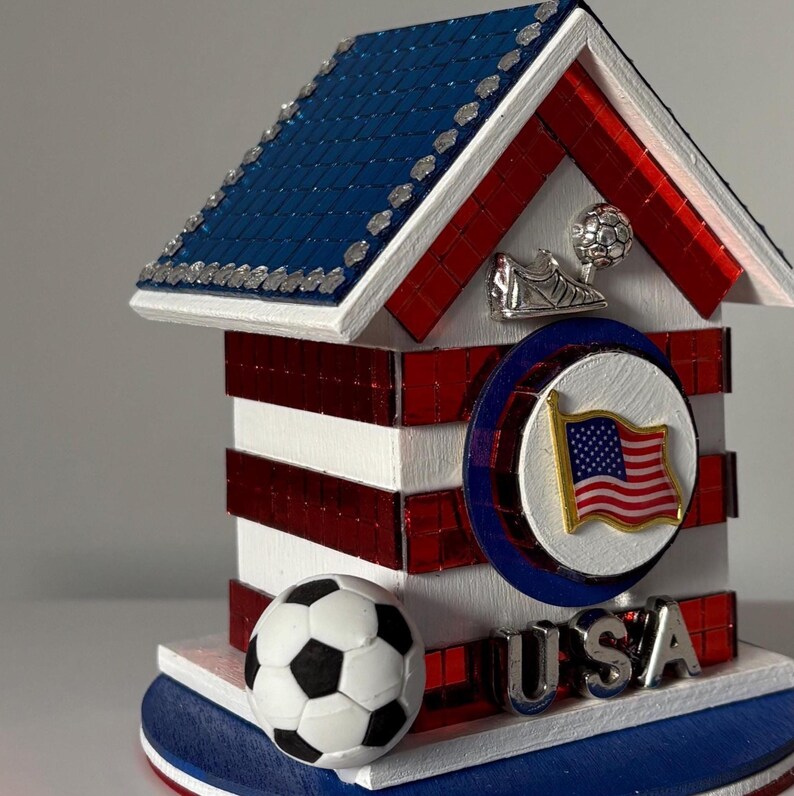 K&ouml;nnte beinhalten: Ein dekoratives patriotisches Haus mit blauem Dach, rot-wei&szlig;en W&auml;nden und einem Fu&szlig;ball. Das Haus zeigt ein USA-Flaggenemblem und das Wort "USA" in silbernen Buchstaben. Ein Fu&szlig;ballschuh und ein Ball sind ebenfalls abgebildet.