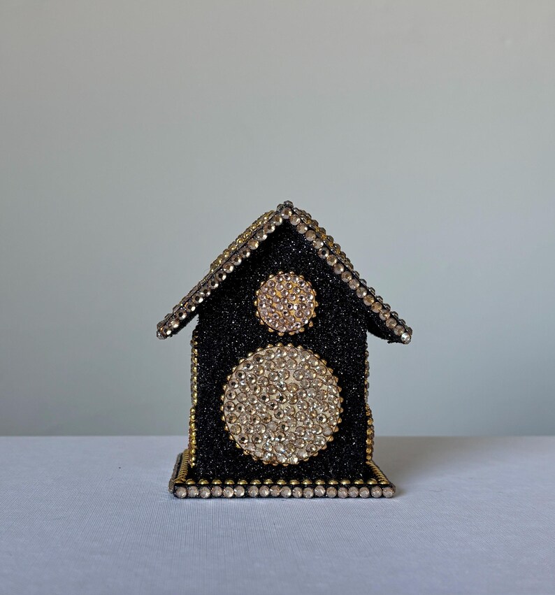 Front view of black glitter rhinestone house ornament with large circular crystal accent and gold trim on white background.