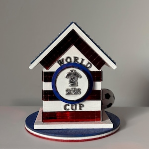 K&ouml;nnte beinhalten: Ein dekoratives Holzhaus in Rot, Wei&szlig; und Blau mit einem Fu&szlig;ball und einem "World Cup 2026" Emblem. Das Haus hat ein gestreiftes Design und eine blaue Basis.