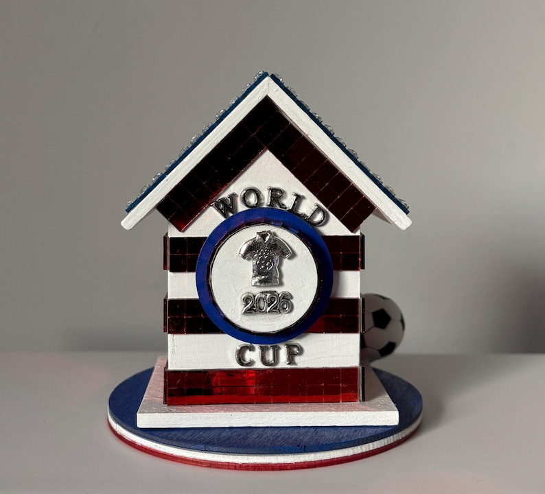 K&ouml;nnte beinhalten: Ein dekoratives Holzhaus in Rot, Wei&szlig; und Blau mit einem Fu&szlig;ball und einem "World Cup 2026" Emblem. Das Haus hat ein gestreiftes Design und eine blaue Basis.