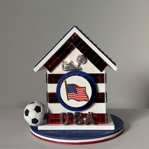 Handcrafted USA soccer themed miniature house with red white and blue mirrored accents, American flag emblem, and small soccer ball on base, shown on a clean neutral background
