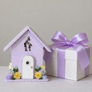 May include: A lavender-colored decorative house with a white arched door, adorned with small white pearls along the roofline and door frame. A silver silhouette of two figures stands above the door, flanked by small yellow and blue paper flowers.