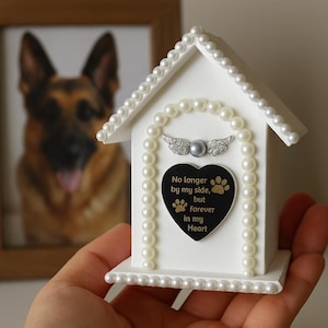 May include: White pet memorial house with pearl detailing and a black heart-shaped plaque. The plaque reads "No longer by my side, but forever in my Heart" with paw prints. A framed photo of a dog is in the background.