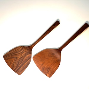 May include: Two wooden spatulas with long, slender handles and wide, angled heads. The spatulas are made of dark brown wood with visible grain patterns, suggesting a natural finish. The kitchen tools are designed for cooking.