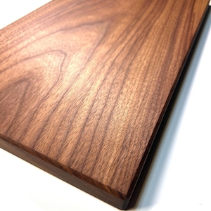 Puede incluir: Una tabla de cortar rectangular de madera de color marrón oscuro, con vetas de madera visibles. La tabla tiene una superficie lisa y pulida y un borde biselado. La madera parece ser de nogal, sobre un fondo blanco.