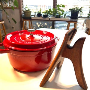 Pode incluir: Uma panela holandesa de ferro fundido esmaltado vermelho vibrante com tampa combinando e botão prateado. Um suporte de madeira está posicionado à direita. A panela está sobre uma superfície branca, sugerindo uma cozinha ou sala de jantar.