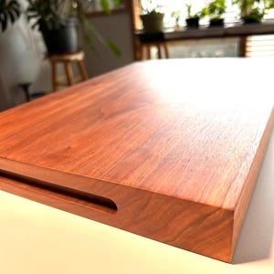 以下が含まれることがあります： 赤褐色がかった豊かな色合いの長方形の無垢材まな板。表面は滑らかで研磨されており、片側にくぼんだ取っ手が付いています。木目がはっきりと見え、自然な美しさを添えています。キッチン用品です。