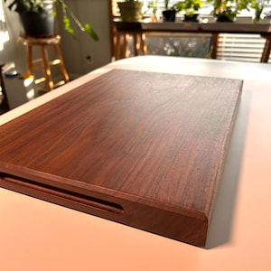 Puede incluir: Una bandeja rectangular de madera para servir, con un acabado marrón oscuro. Tiene un asa empotrada en un lado y se apoya sobre una superficie clara. La veta de la madera es visible, lo que añade a su estética natural.