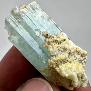May include: A pale blue aquamarine crystal with a rough, textured surface. The crystal is partially embedded in a beige matrix of rock and minerals. The aquamarine has a translucent quality, with visible striations.