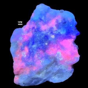 May include: A glowing, irregularly shaped mineral specimen with a black background. The mineral displays a vibrant color palette of pink, blue, and white, with the text "Long wave" in the upper left corner. The colors are concentrated in a cloud-like pattern.